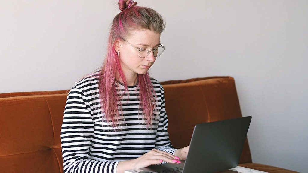 Une jeune femme avec une mèche de cheveux rose est assise sur un canapé et travaille sur un ordinateur portable. 