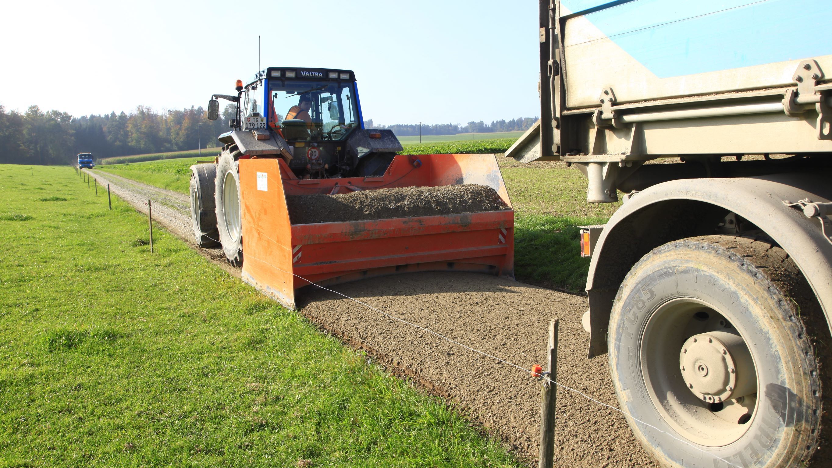 Ein Grader mit orangefarbenem Schild und ein LKW bringen eine neue Schotterschicht auf. Der Schotterweg führt durch eine Wiese.