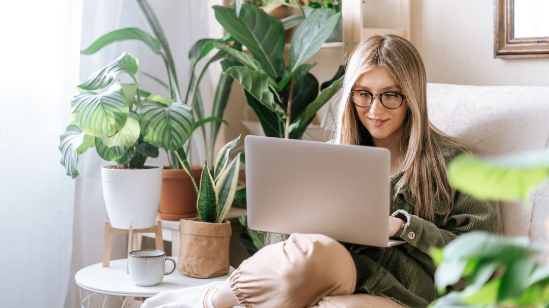 A woman is sitting in an armchair with a laptop on her lap, surrounded by plants.