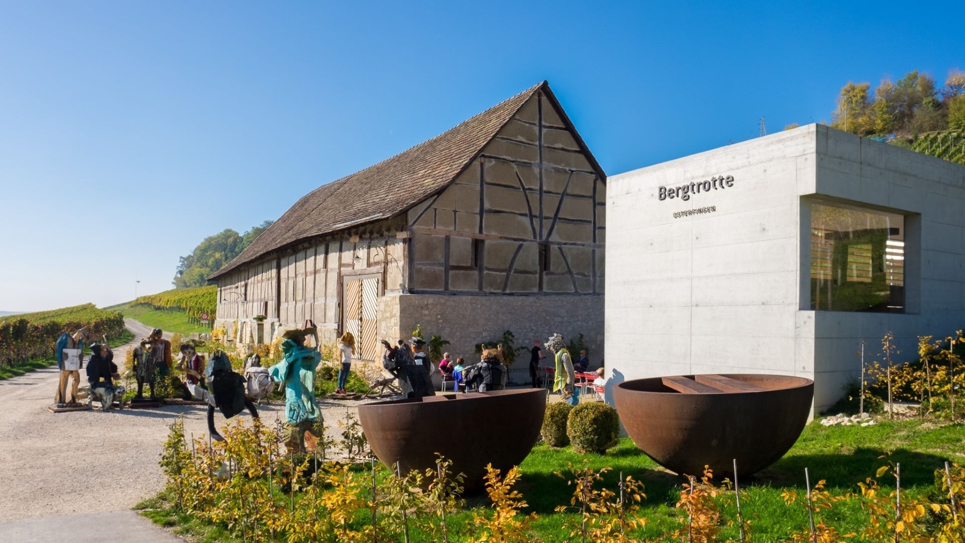La Bergtrotte Osterfingen, un vénérable bâtiment à colombages avec un imposant toit en bâtière, est située au milieu du vignoble aux couleurs automnales. Elle est complétée par une annexe moderne en béton, de forme carrée, sur le côté frontal droit. Entre les deux se trouve une petite cour intérieure. Différents objets d'art sont placés le long des deux bâtiments. Outre des figures en bois peintes en couleur, on peut voir deux grandes demi-coques en acier rouillé. Le ciel est bleu et sans nuages.