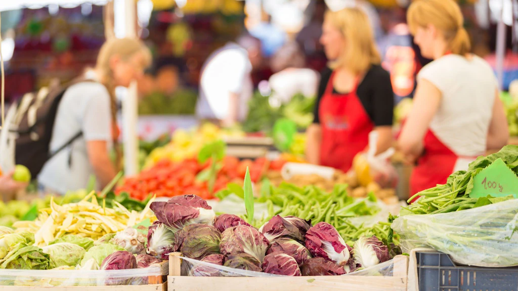 Sur un marché, différentes sortes de salades et de légumes sont présentées dans des caisses en bois. À l’arrière-plan, on voit deux vendeuses et une cliente portant un sac à dos.