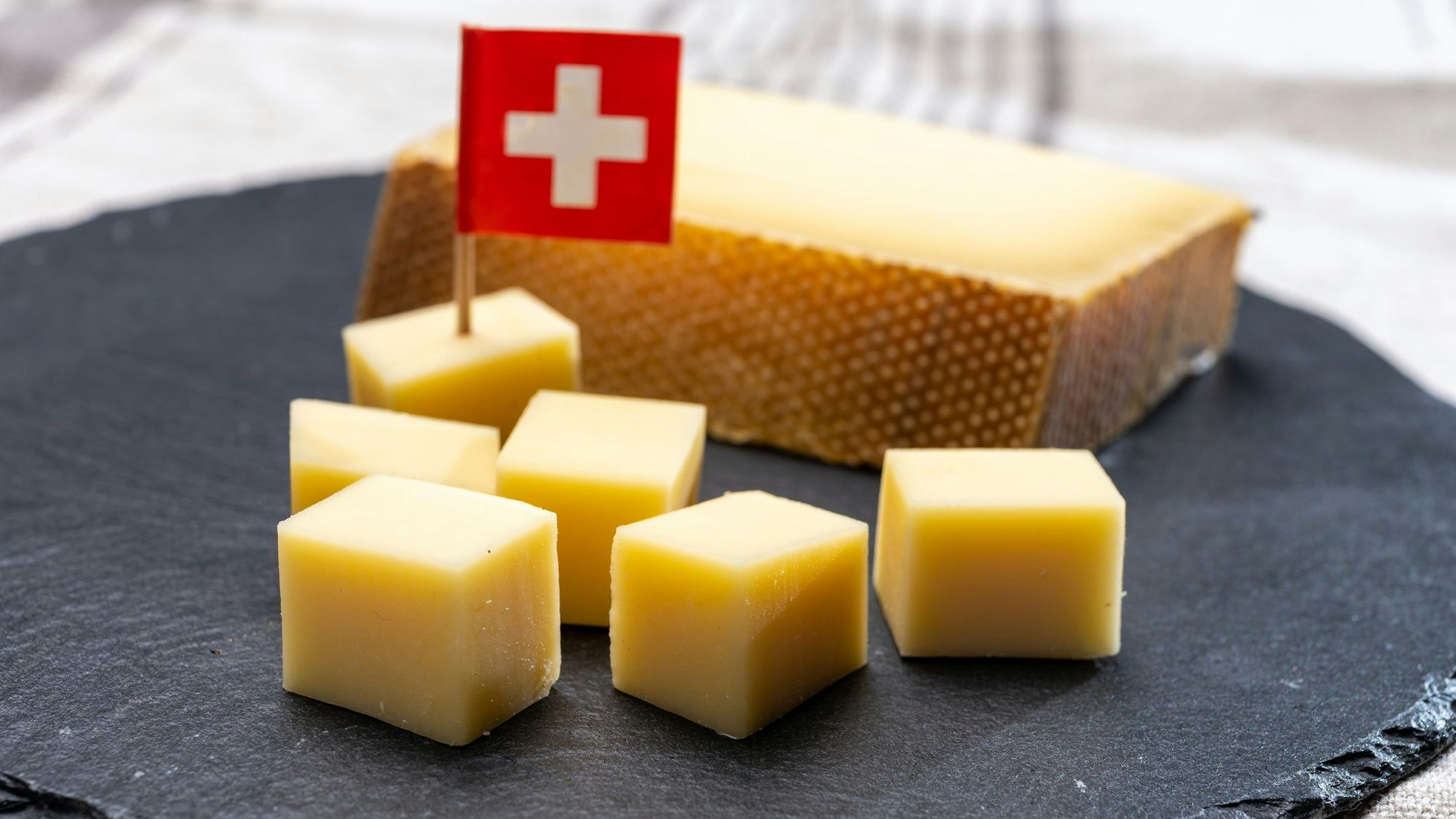Un fromage suisse présenté en cubes sur une plaque d’ardoise : au-dessus de l’ensemble apparaît un petit drapeau suisse.