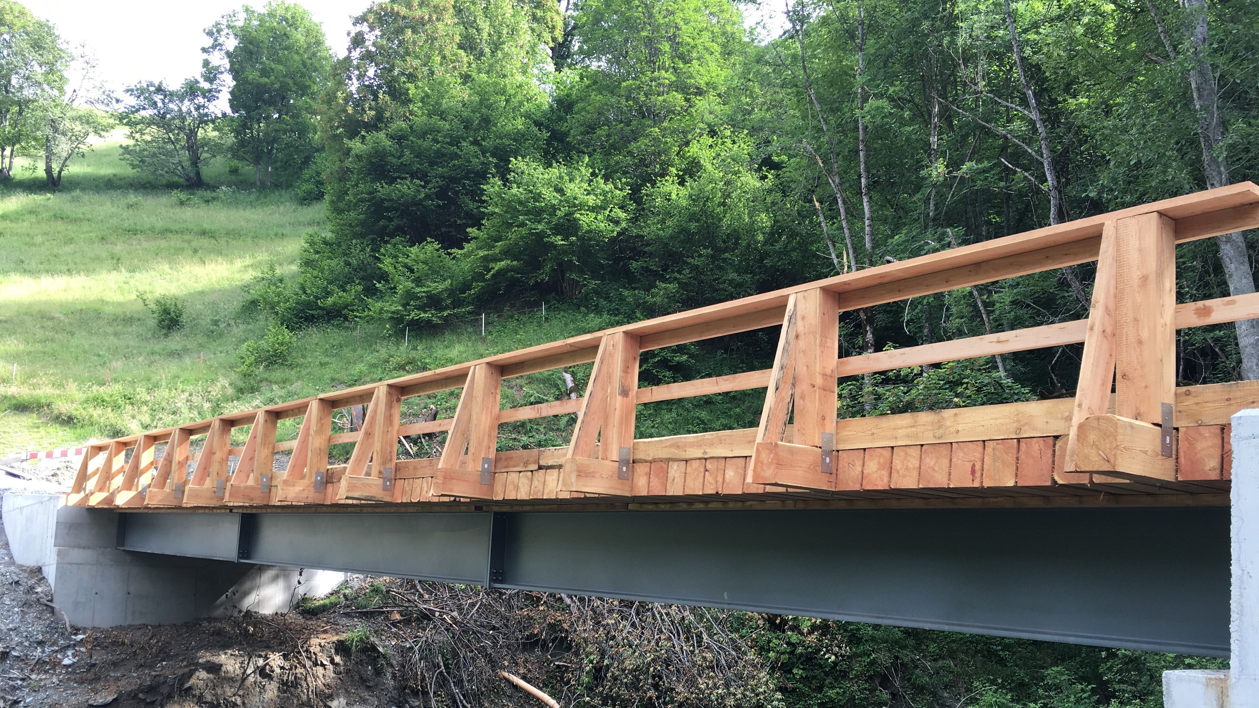 Un nuovo ponte in legno con ringhiera in legno sovrasta un torrente. Gli appoggi sono in calcestruzzo e l’impalcato è costituito da una trave in acciaio.