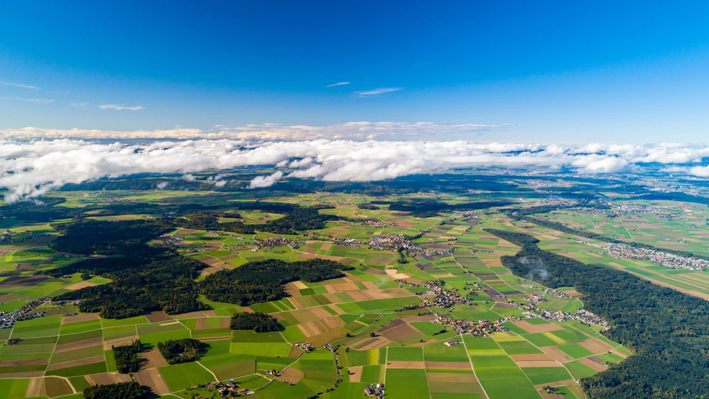 Veduta aerea di un terreno agricolo svizzero con campi verdi e marroni circondati da foreste. Piccoli villaggi sono sparsi nel paesaggio, mentre una coltre di nuvole aleggia sopra di essi. L'immagine simboleggia l'interazione tra ambiente, questioni sociali e uso economico.