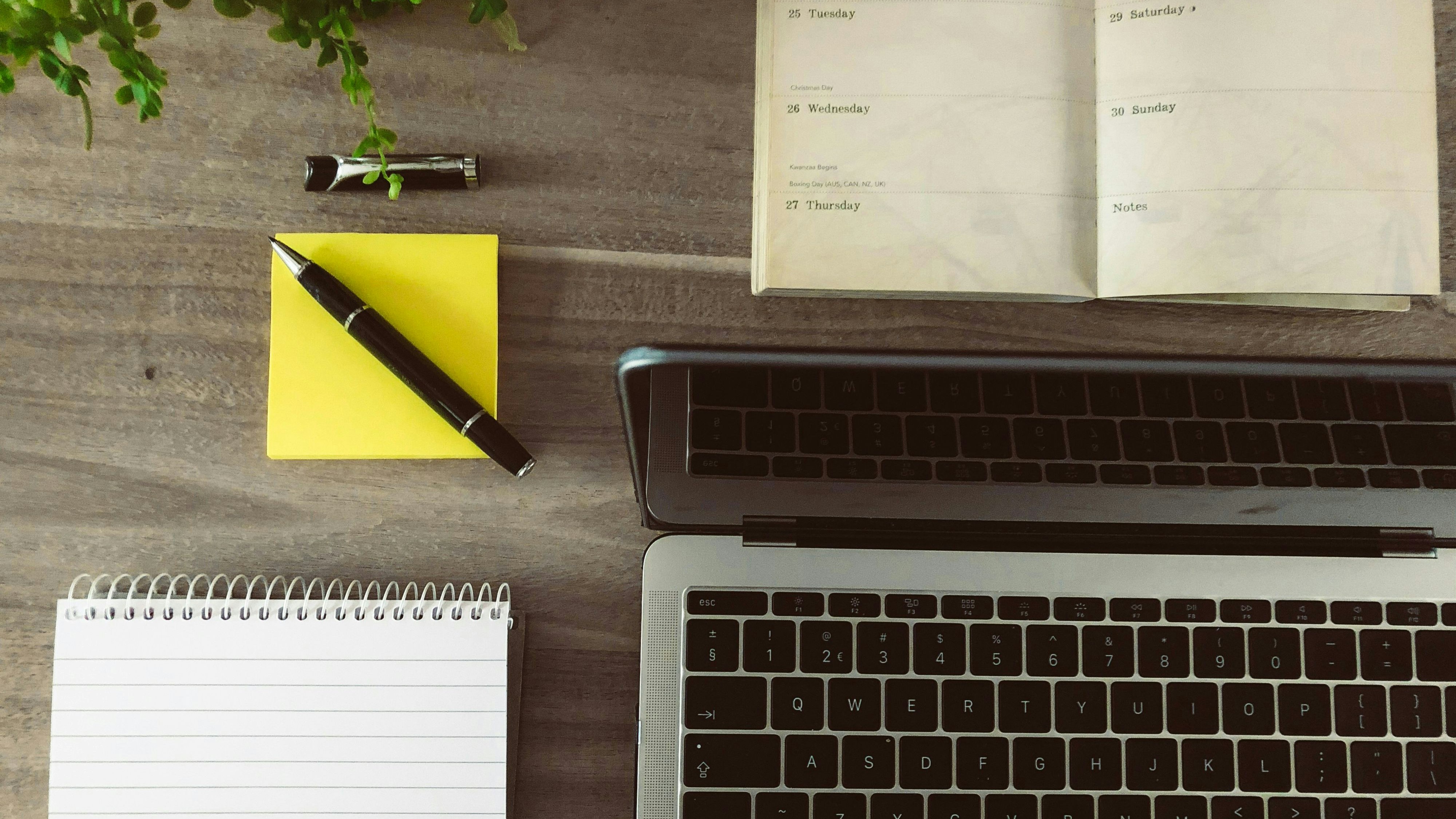 Un blocco note giallo con penna a sfera, agenda e laptop visti dall'alto.