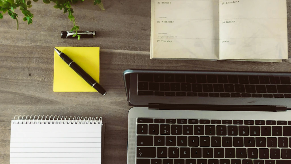 Un blocco note giallo con penna a sfera, agenda e laptop visti dall'alto.